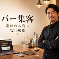 Webサイトを表示したノートパソコンとスマートフォンを前に、店内で腕を組む日本人バーテンダー
